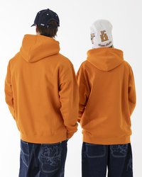 Two individuals facing away, wearing orange hoodies. One has a navy cap, the other a white beanie with text. Denim pants feature patterns.