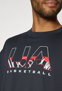 Mørkegrå t-shirt med "UA BASKETBALL" i store hvide og røde bogstaver med et tekstureret mønster, der ligner et basketballnet.