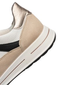 Detailní záběr paty tenisky s béžovou semišovou částí, bílou texturovanou kůží, černým detailem, kovovým zlatým lemováním a bílou pryžovou podrážkou s vzorkem.