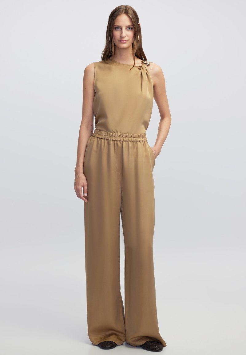 Tuta color cammello con design senza maniche, dotato di dettaglio a nodo sulla spalla, cintura elastica e pantaloni a gamba larga. Tessuto a superficie liscia.