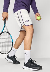 Valged tennisepüksid, millel on lilla ääris ja Adidas logo, koos mustade spordijalatsitega ja valgete sokkidega, hoides tennisereketit ja -palli.