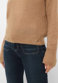 Bruine wollen trui met een zachte textuur, opgerolde manchetten en een klein accentdetail aan de zoom, gecombineerd met donkerblauwe jeans met achterzakken.