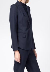 Námořnický modrý přizpůsobený blazer, zapínání na jeden knoflík, klopové límce, dvě přední kapsy, hladká textura, dlouhé rukávy s knoflíkovými akcenty.