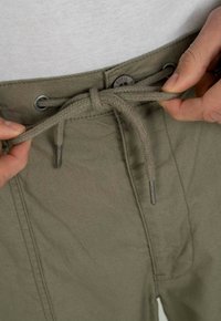 Olivgrüne Hose mit Kordelzugbund, ausgestattet mit Metall-Ösen und einem strukturierten Stoff. Nahaufnahme des Bundbereichs mit Händen, die den Kordelzug halten.