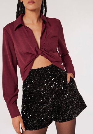 Burgunderfarbendes Strickshirt mit einem Knoten-Detail vorne, kombiniert mit schwarzen, mit Pailletten besetzten Shorts, die eine weiche Textur und eine lockere Passform haben.