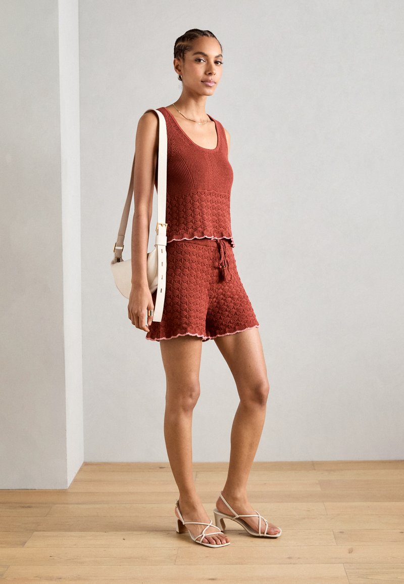 Rostfarbenes gestricktes Tanktop mit gewelltem Saum, kombiniert mit passenden Shorts. Das Model trägt eine weiße Umhängetasche und zieht Riemchensandalen an.