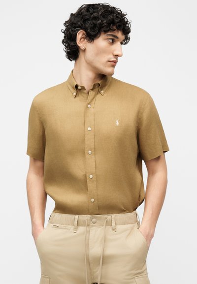 Άνδρας με σγουρά σκούρα μαλλιά φοράει κοντομάνικο πουκάμισο ταμπά, φορεμένο μέσα σε ανοιχτό μπεζ παντελόνι, τα χέρια στις τσέπες, κοιτάει πλάγια.