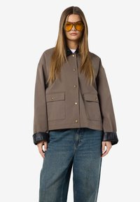 Jonge vrouw in een oversized bruine jas met zwarte manchetten, grote voorkruim pockets, blauwe jeans en gele piloten zonnebril, rechtop staand.