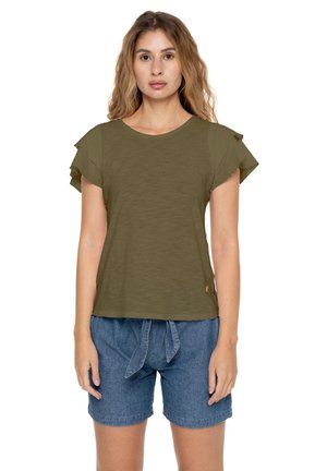 Une femme porte un haut vert olive à manches volantées et un short en jean bleu sur fond blanc.
