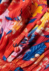 Tessuto floreale rosso con accenti blu e gialli; presenta una superficie texturizzata e bottoni rotondi lungo il patta anteriore.