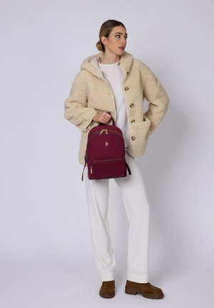 Giacca beige in finta pelliccia, zaino bordeaux con zip dorata, outfit bianco, stivali marroni con texture, modella in piedi contro uno sfondo neutro.