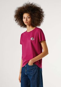 Fuksijos spalvos marškinėliai su apvalia iškirpte, pagaminti iš minkštos medžiagos, su sidabriniu lūpų atspaudu kairiajame krūtinės kampe. Laisva forma, trumpomis rankovėmis, lygi tekstūra.