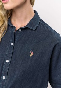 Donker denim overhemd met een kraag, voorzien van een knoopsluiting en een klein geborduurd logo op de linkerborst. Gladde textuur, klassieke pasvorm.