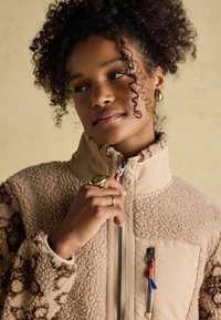 Young woman with curly hair wearing a beige fleece jacket, gold earrings, and rings, holding the jacket zipper near her chin.