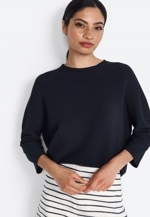 Marineblauer Strickpullover mit rundem Ausschnitt und drei Viertelärmeln. Kombiniert mit einem gestreiften Rock in Weiß und Marineblau. Glatte Textur.