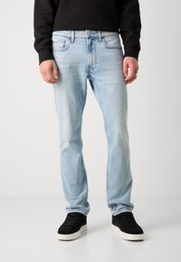 Hellblaue Jeans mit geradem Bein und ausgewaschenem Finish, ausgestattet mit fünf Taschen und einem Metallknopfverschluss. Kombiniert mit schwarzen Turnschuhen.