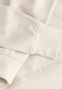 Gros plan des poignets et de l'ourlet beige clair d’un sweat-shirt avec des coutures apparentes sur un tissu en coton doux.