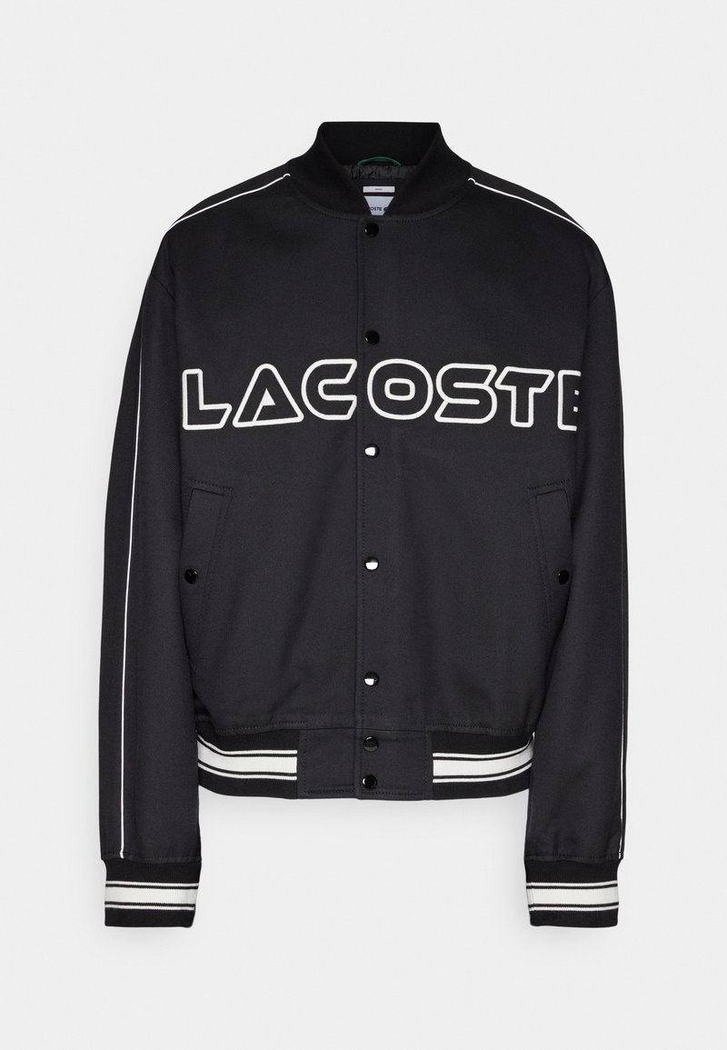 Veste bomber noire avec un logo « LACOSTE » blanc prominent, des boutons-pression, des poches latérales et des accents rayés sur les poignets et l'ourlet.