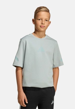 Niño pequeño con cabello rubio corto que lleva una camiseta holgada de color gris claro y pantalones negros con logotipos de Adidas, de pie frente a un fondo liso.