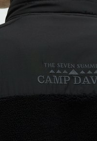 Dos d'une veste polaire noire avec le texte brodé "THE SEVEN SUMMITS" et "CAMP DAVID" sous de petites icônes de montagnes.