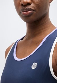 Κοντινό πλάνο ενός ατόμου που φοράει μπλε ναυτικό αμάνικο τοπ με λευκή και λιλά λεπτομέρεια, με μικρό λογότυπο K-Swiss στο στήθος.