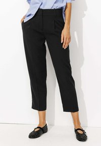 Pantalons noirs courts avec des plis à l'avant, fabriqués en tissu lisse. Associés à des chaussures noires plates avec des clous argentés et un bout rond.