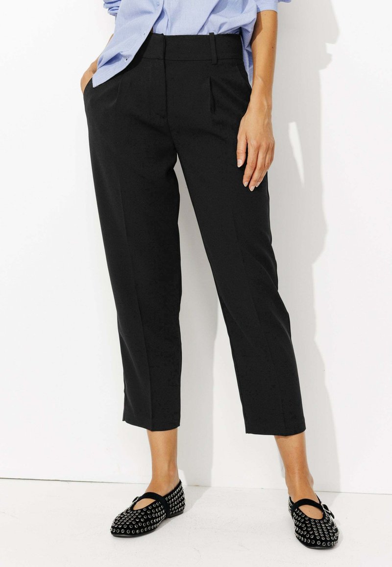 Pantalons noirs courts avec des plis à l'avant, fabriqués en tissu lisse. Associés à des chaussures noires plates avec des clous argentés et un bout rond.