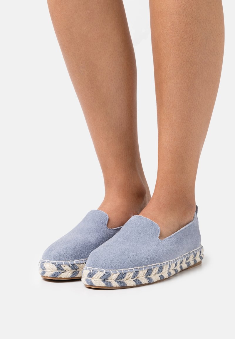 Pieds chaussés d'espadrilles légères en daim bleu clair à enfiler, avec une semelle en jute tressé et des détails blancs, sur un fond blanc uni.
