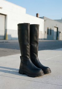 Cizme negre înalte din piele, cu tălpi groase, stând în picioare pe o trotuar de beton într-un mediu urban.