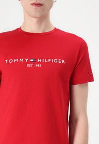 Camiseta de algodón roja con un logo blanco que dice "TOMMY HILFIGER EST. 1985" y un pequeño emblema en la manga. Mangas cortas, cuello redondo.
