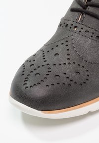Chaussure en cuir noir avec un design perforé, bout arrondi et semelle en caoutchouc blanc. Présente des motifs délicats pour la ventilation et le contraste de texture.