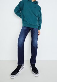 Turkusowa bluza z kangurową kieszenią, niebieskie dżinsy, czarne i szare trampki za kostkę, na jednolitym białym tle.