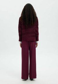 Bordo spalvos tekstūruotas megztinis ir plačios kelnės, pasiūtos tiesaus kirpimo su subtiliais raštais. Matomos ilgos garbanotos plaukai, modelis žiūri nugara.