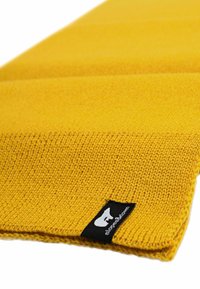 Coperta gialla lavorata a maglia con un motivo testurizzato e un'etichetta nera cucita che presenta un logo. Il tessuto appare morbido e caldo.
