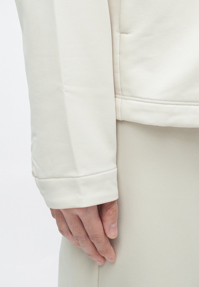 Main reposant détendue sur le côté, portant un vêtement à manches longues beige clair avec des coutures visibles et un tissu texturé.