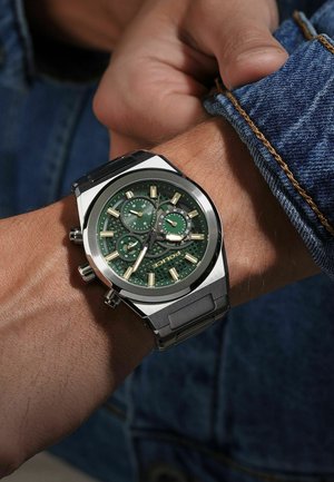 Orologio in acciaio inox satinato argentato con quadrante verde texturizzato, indici delle ore dorati, tre sub-quadranti e cinturino in metallo, indossato su un polso con jeans.