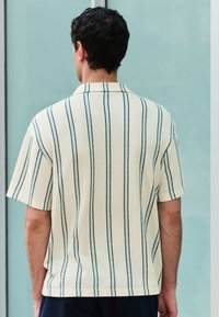 Short-sleeve, collar shirt in cream with vertical dark green stripes. Textured fabric with a relaxed fit, visible stitching along edges.
