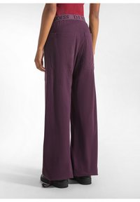Bordeaux wijd-uitlopende broek met een verstelbare tailleband met de tekst "GODDESS". Twee achterzakken en een zachte, gestructureerde stof. Zwarte schoeisel zichtbaar.