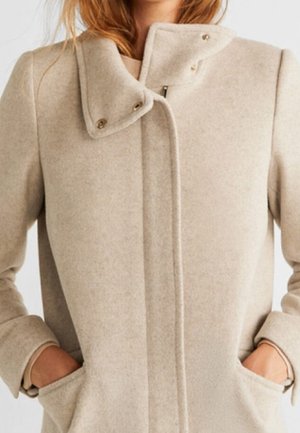 Femme portant un manteau en laine beige avec col à pressions, fermeture éclair dissimulée et les mains dans les poches avant, sur fond clair.