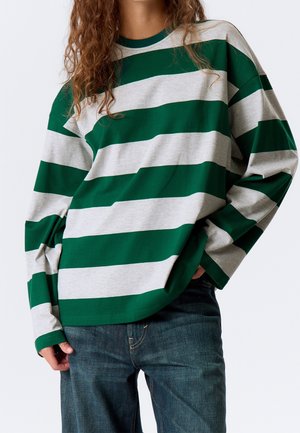 Person trägt ein langärmeliges Hemd mit breiten grünen und hellgrauen horizontalen Streifen und dunkelblaue Jeans, Hand in der Tasche.