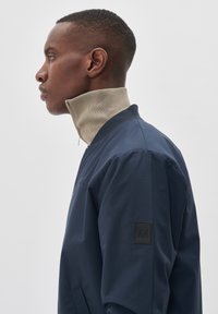 Marineblauwe bomberjack met een beige geribbelde kraag, ritssluiting en een getextureerd logo op de linkermouw. Getailleerde pasvorm, gladde stof.