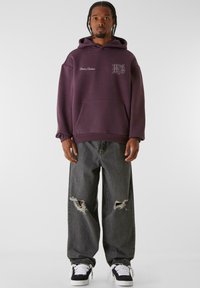 Lilla hettegenser med frontlomme, kombinert med slitte grå jeans med hull. Svarte joggesko med hvite såler fullfører antrekket.