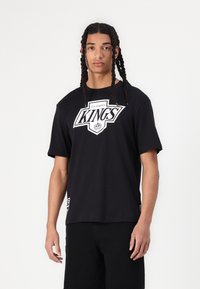 Crna pamučna majica s velikim bijelim logotipom "Los Angeles KINGS", kratkim rukavima i opuštenim krojem. Sadrži oznaku brenda sa strane.