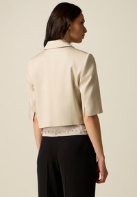 Mujer con un blazer crema corto de mangas hasta el codo sobre una blusa adornada con perlas y pantalones negros, mirando de perfil contra un fondo liso.