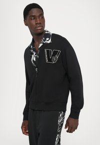 Melna zip-up svītra ar V88 logo uz krūtīm un rakstainu krekla apkakli. Iespējas ietver ribotas piedurknes un strukturētu audumu.