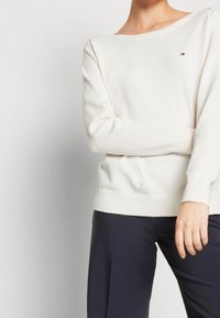 Pull beige clair avec une texture douce et un large col, portant un petit logo sur la poitrine. Poignets et ourlet côtelés.