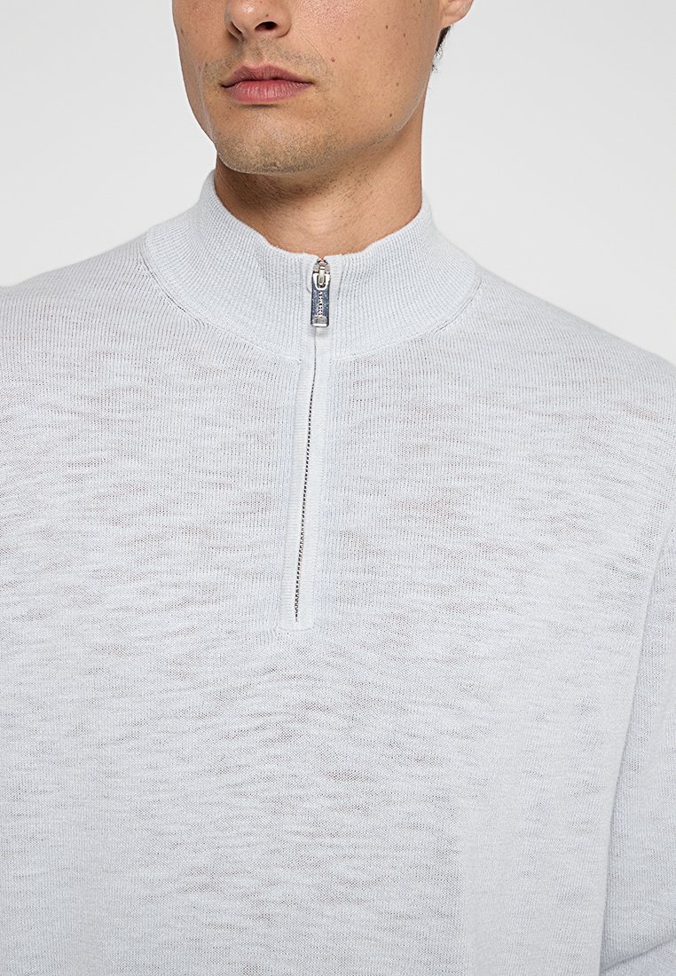 Hellgrauer Strickpullover mit hohem Kragen und halblangem Reißverschluss. Besitzt ein strukturiertes Muster und subtile Variationen in der Garnstärke.
