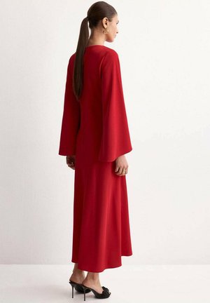 Mujer con una larga coleta, vestida con un vestido largo rojo de mangas anchas y zapatos negros de tacón alto y puntera abierta, de pie de perfil sobre un fondo liso.