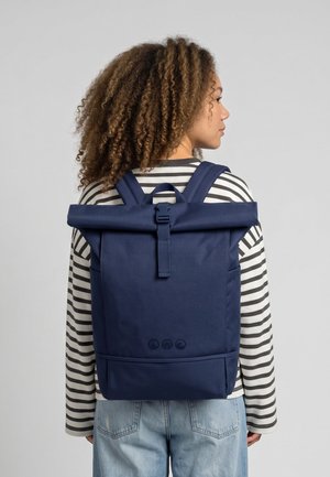 Frau mit lockigem Haar, die ein gestreiftes Hemd und hellblaue Jeans trägt und einen großen dunkelblauen Rolltop-Rucksack trägt.