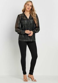 Blusa nera trasparente con strisce verticali argento, scollatura a V e maniche a palloncino, abbinata a jeans skinny neri e tacchi metallici.